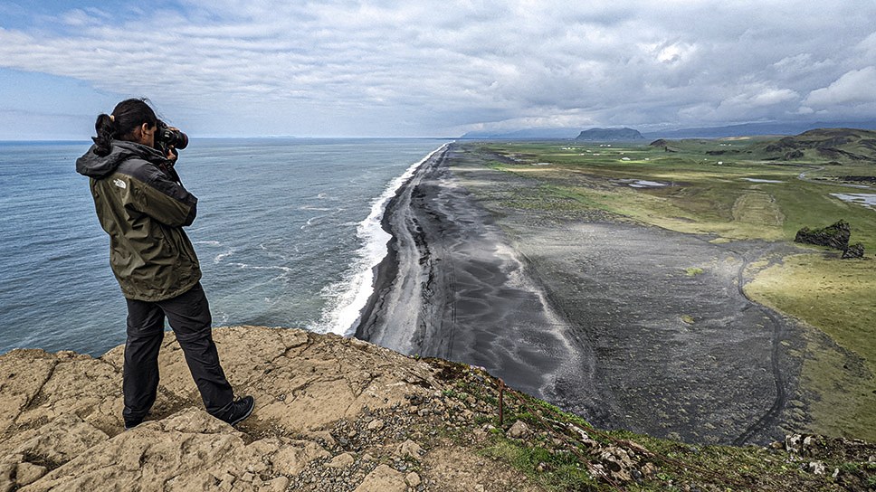 Fotograf mit Kamera auf einer Klippe über Meer und schwarzem Strand in Island